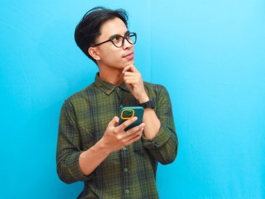 Ciddi Asyalı bir adam elinde cep telefonu tutarken mavi arka plana bakarak düşünme hareketi yapıyor.