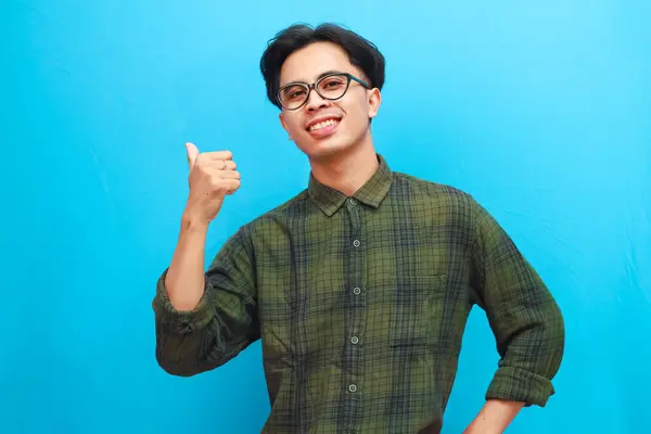 stock image Portrait smiling young Asian man pointing to the side with thumb on blue isolated studio background.