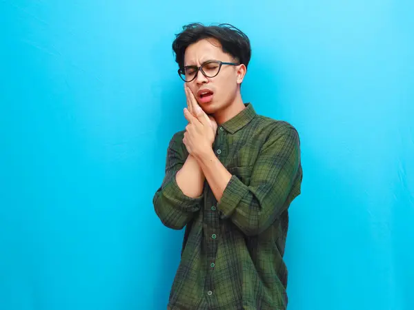 stock image Portrait of unhealthy Asian man posing with toothache and holding right cheek with pain expression on blue background.