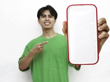 Yeşil tişört giyen gülümseyen genç Asyalı bir adamın portresi, elinde akıllı bir telefon ve beyaz bir arka planda izole edilmiş büyük boş bir ekran gösteriyor..