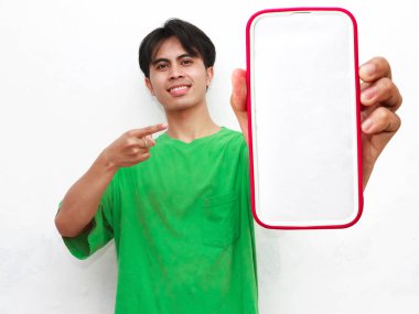 Yeşil tişört giyen gülümseyen genç Asyalı bir adamın portresi, elinde akıllı bir telefon ve beyaz bir arka planda izole edilmiş büyük boş bir ekran gösteriyor..