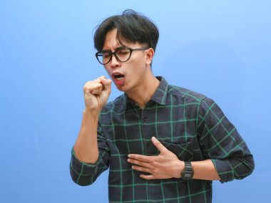 Hüzünlü ve üzgün bir ifadeyle öksüren sağlıksız bir Asyalı adamın portresi mavi arka planda izole edilmiş..