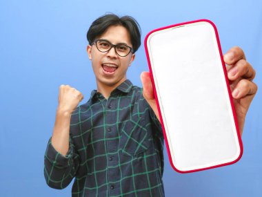 Boş bir akıllı telefon ekranı ve sıkılmış bir yumrukla poz veren Asyalı bir adamın portresi zaferi mutlu, gülümseyen bir ifadeyle kutluyor. Mobil teklifleri, uygulamaları veya reklamları göstermek için ideal.