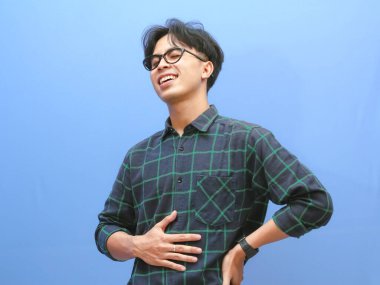 Portrait of a smiling Asian man in a casual shirt, touching his belly with a relieved expression isolated against a blue background. clipart