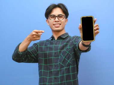 Asyalı genç adam sarı kazak giyiyor ve akıllı telefonu işaret ediyor.