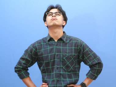 portrait of a stressed young adult Asian man wearing glasses holding his head with a sad expression isolated on a blue background clipart