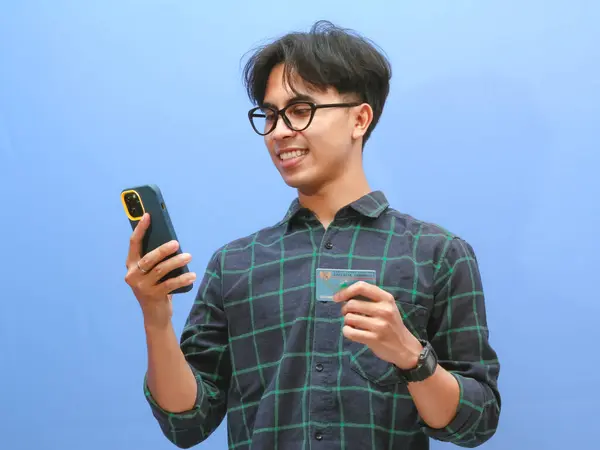 stock image Portrait of a happy Indonesian man in a casual shirt smiling while using a mobile phone and holding an ID card or KTP, looking directly at the cameraisolated against a blue background.