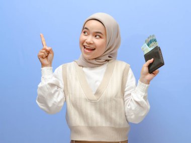 Para dolu bir cüzdan taşıyan heyecanlı bir Asyalı kadının portresi, mutlu bir jest gösteriyor, mavi bir arka planda izole edilmiş..