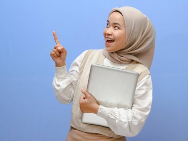 Endonezyalı güzel bir kız öğrencinin portresi dizüstü bilgisayarını tutarken heyecanlı bir ifadeyle yukarı bakıyor, mavi bir arkaplana karşı bir fikri işaret ediyor..