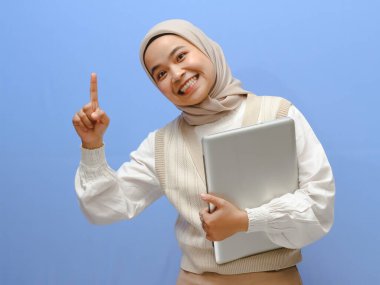Endonezyalı güzel bir kız öğrencinin portresi dizüstü bilgisayarını tutarken heyecanlı bir ifadeyle yukarı bakıyor, mavi bir arkaplana karşı bir fikri işaret ediyor..