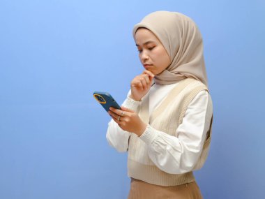 Ciddi Asyalı Müslüman kadın mavi arka planda cep telefonuna bakarken derin düşüncelere dalmış.