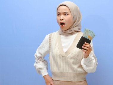 Şok olmuş bir ifadeyle Asyalı bir kadının portresi. Elinde para dolu bir cüzdan, şaşkınlıkla bekliyor. Mavi arkaplan.