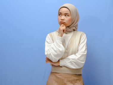 Asyalı ciddi Müslüman bir kadının portresi mavi bir arka planda el pençe içinde düşünme hareketi gösteriyor..