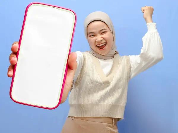 stock image Portrait of an asian woman holding a smartphone, showing and presenting a blank screen mobile phone, clenching her fists, celebrating victory with a happy and excited expression.