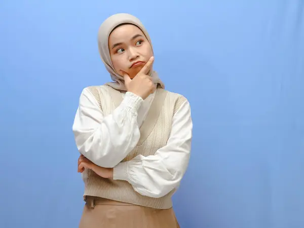 stock image Portrait of serious asian muslim woman showing thinking gesture with hand in chin isolated on a blue background.