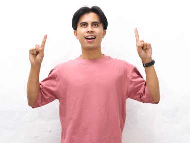 Pembe tişörtlü yakışıklı Asyalı genç bir adam yukarı doğru parmak pozu veriyor, beyaz arka plana karşı hayret verici bir ifade sergiliyor..