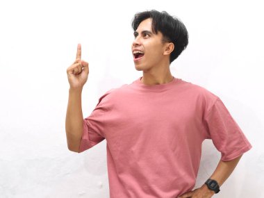 Pembe tişörtlü yakışıklı Asyalı genç bir adam yukarı doğru parmak pozu veriyor, beyaz arka plana karşı hayret verici bir ifade sergiliyor..