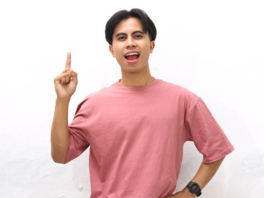 Pembe tişörtlü yakışıklı Asyalı genç bir adam yukarı doğru parmak pozu veriyor, beyaz arka plana karşı hayret verici bir ifade sergiliyor..