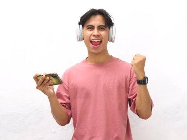 Pembe tişörtlü heyecanlı genç Asyalı adam, kablosuz kulaklık takıyor video oyunu oynuyor ya da bir dereyi seyrediyor zaferi ve başarıyı beyaz arka planda izole edilmiş gururlu bir çığlıkla kutluyor.