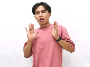 Pembe tişörtlü genç Asyalı adam beyaz bir arka planda panik ifadesiyle şoka girmiş görünüyor..