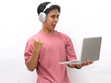 Pembe tişörtlü genç Asyalı adam elinde kablosuz kulaklıkla dizüstü bilgisayarla gururla bağırıyor ve zaferi kutluyor beyaz bir arka plana karşı heyecanlı bir ifadeyle.