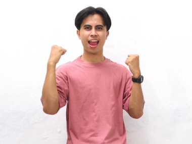 Pembe tişörtlü, kendine güvenen genç Asyalı adam yumruklarını sıkmış ve beyaz bir arka plana karşı heyecanlı bir ifadeyle poz veriyor..