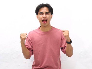 Pembe tişörtlü, kendine güvenen genç Asyalı adam yumruklarını sıkmış ve beyaz bir arka plana karşı heyecanlı bir ifadeyle poz veriyor..