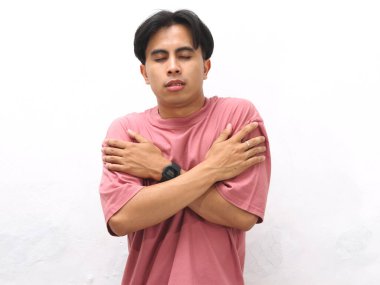 Pembe tişörtlü sağlıksız genç Asyalı adam acı ve üzüntü hissediyor. Üşüyormuş gibi kendine sarılıyor ya da beyaz arka planda titriyor..