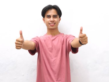 Pembe tişörtlü, kendine güveni tam bir Asyalı. Başparmakları havada poz veriyor ve kameraya beyaz arka planda gülümsüyor..