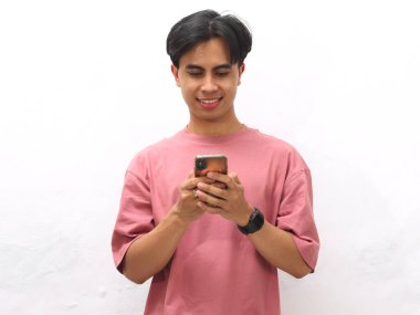 Pembe tişörtlü genç Asyalı adam, beyaz bir arka planda cep telefonunu tutarken gülümsüyor ve odaklanıyor..
