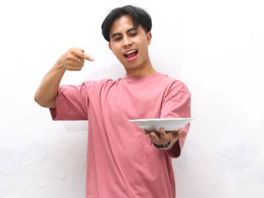 Pembe tişörtlü genç Asyalı bir adam elinde boş bir tabakla beyaz bir arkaplan karşısında şaşkın ve heyecanlı bir ifadeyle işaret ediyor..