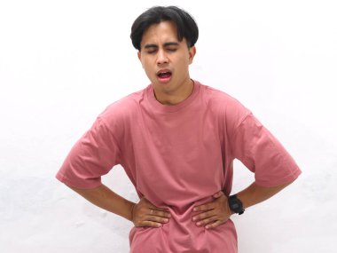 Pembe tişörtlü sağlıksız genç Asyalı adam beyaz bir arka planda acı ve üzüntü içinde karnını tutuyor..