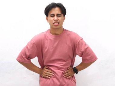 Pembe tişörtlü sağlıksız genç Asyalı adam beyaz bir arka planda acı ve üzüntü içinde karnını tutuyor..