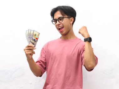 Pembe tişörtlü genç bir Asyalı 'nın portresi nakit para taşıyor, zaferi bir yumrukla kutluyor ve beyaz izole edilmiş bir arka plana karşı mutlu, heyecanlı bir yüz ifadesi takınıyor..