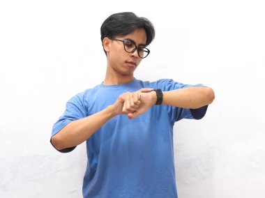 Mavi gömlekli genç bir Asyalı siyah saatini beyaz arka planda kontrol ederken endişeli görünüyor..