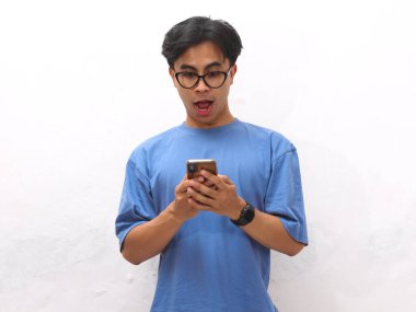 Mavi tişörtlü genç Asyalı adamın portresi. Komik bir yüz ifadesi var. Asyalı adam elinde akıllı bir telefon tutuyor..