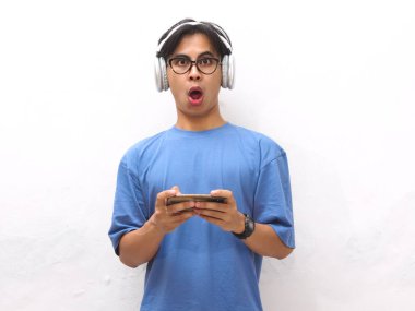 Mavi tişörtlü genç Asyalı adam kablosuz kulaklık kullanıyor, video oyunu oynuyor ya da beyaz bir arka planda izole edilmiş komik bir suratla akıllı bir telefondan akarsu izliyor..