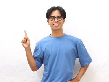 Young Asian man in a blue t-shirt pointing at blank copy space, ideal for promotional or advertising content. clipart