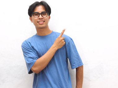 Young Asian man in a blue t-shirt pointing at blank copy space, ideal for promotional or advertising content. clipart