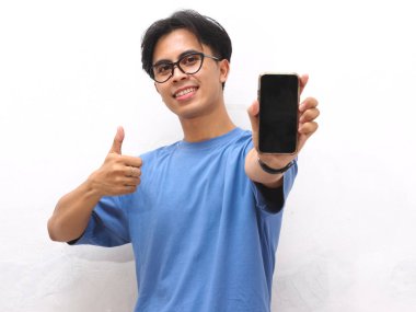 Mavi tişörtlü genç Asyalı adam gülümsüyor, tamam işareti yapıyor ve boş bir telefon ekranı tutarken baş parmak işareti yapıyor. Beyaz arka planda boş ekranlı telefon gösteren yakışıklı Asyalı genç adam..