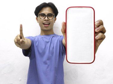 Mavi tişörtlü gülümseyen Asyalı bir adam tek parmağıyla işaret ediyor ve elinde boş bir telefon ekranı tutuyor. Beyaz arka planda boş ekranlı telefon gösteren yakışıklı bir Asyalı..