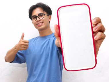 Mavi tişörtlü genç Asyalı adam gülümsüyor, tamam işareti yapıyor ve boş bir telefon ekranı tutarken baş parmak işareti yapıyor. Beyaz arka planda boş ekranlı telefon gösteren yakışıklı Asyalı genç adam..