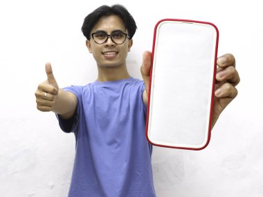 Mavi tişörtlü genç Asyalı adam gülümsüyor, tamam işareti yapıyor ve boş bir telefon ekranı tutarken baş parmak işareti yapıyor. Beyaz arka planda boş ekranlı telefon gösteren yakışıklı Asyalı genç adam..