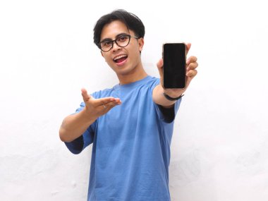 Mavi tişörtlü genç Asyalı adam boş bir telefon ekranını gösterirken gülümsüyor. Beyaz arka planda boş ekran telefon gösteren yakışıklı bir Asyalı..