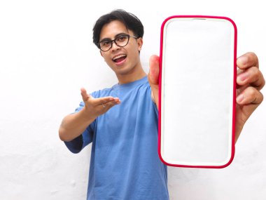 Young Asian man in a blue t-shirt smiling presenting a blank phone screen while pointing,handsome young asian man showing blank screen phone isolated on a white background. clipart