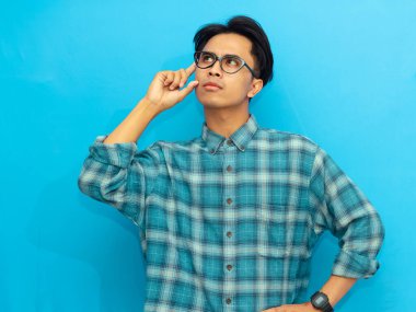 Mavi günlük gömlekli Asyalı bir adam ciddi düşünen bir ifadeyle poz veriyor, mavi arka plana odaklanmış gözlerle bakıyor..