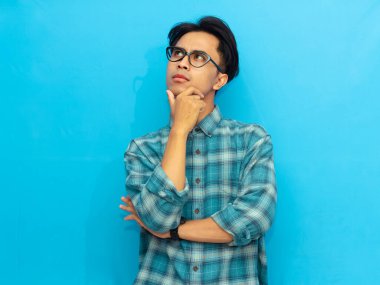 Mavi günlük gömlekli Asyalı bir adam ciddi düşünen bir ifadeyle poz veriyor, mavi arka plana odaklanmış gözlerle bakıyor..