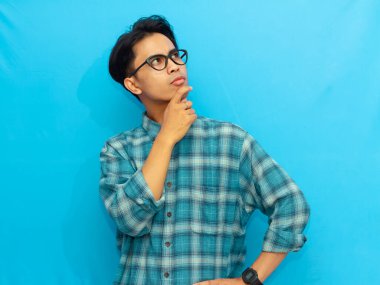 Mavi günlük gömlekli Asyalı bir adam ciddi düşünen bir ifadeyle poz veriyor, mavi arka plana odaklanmış gözlerle bakıyor..