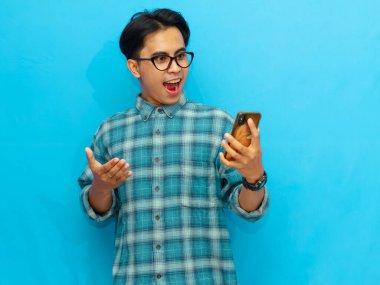 Mavi gömlekli heyecanlı bir Asyalı adam mavi arka planda izole edilmiş sürpriz bir ifadeye sahip bir cep telefonuna bakıyor..
