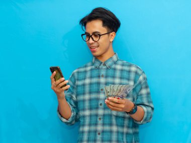 Mavi tişörtlü heyecanlı Asyalı adam elinde cep telefonu ve parayla mavi arka planda izole edilmiş mutlu bir yüz ifadesi gösteriyor..